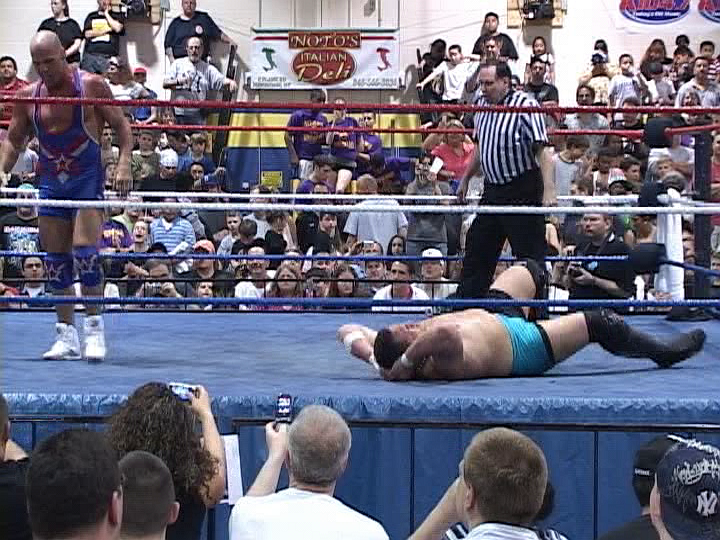 A wrestler stands to the left, a second wrestler on the mat. Referee Dwinell standing to the right.