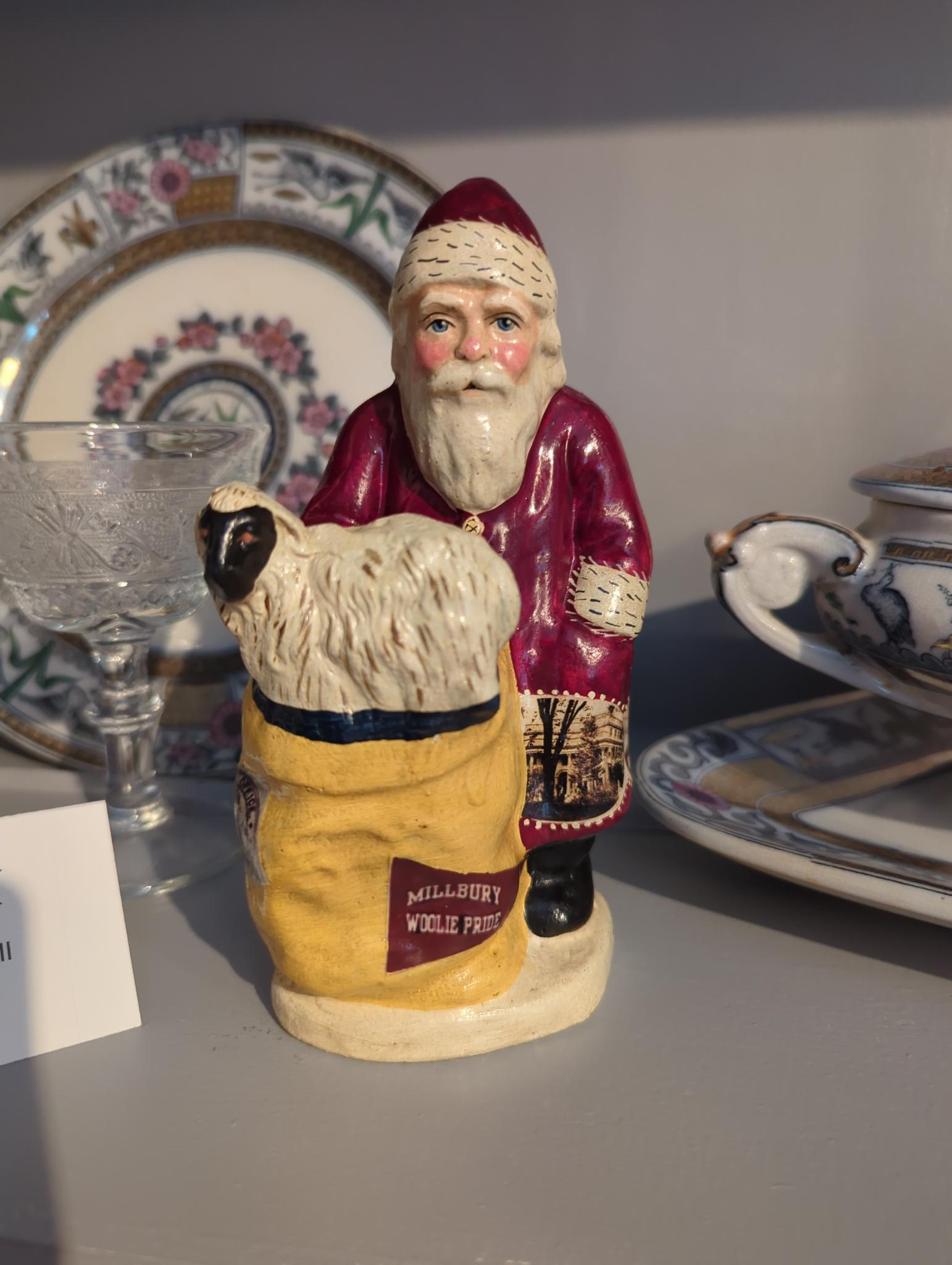 A Santa Claus figurine stands behind a yellow sack with a sheep coming out of it, representing "Millbury Woolie Pride." He is surrounded by pieces of china dinnerware, he wears a red jacket with a painted-on sewn image of the Asa Waters Mansion.