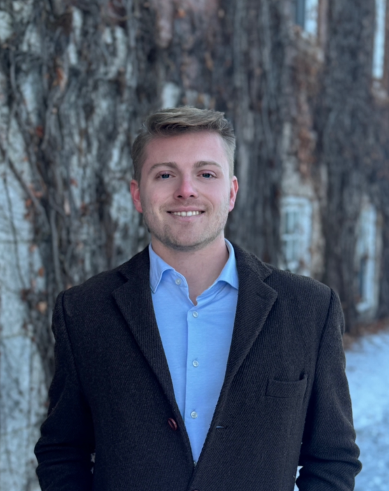A young man stands in front of an ivy-draped wall in a suit jacket and button down blue shirt, no tie.