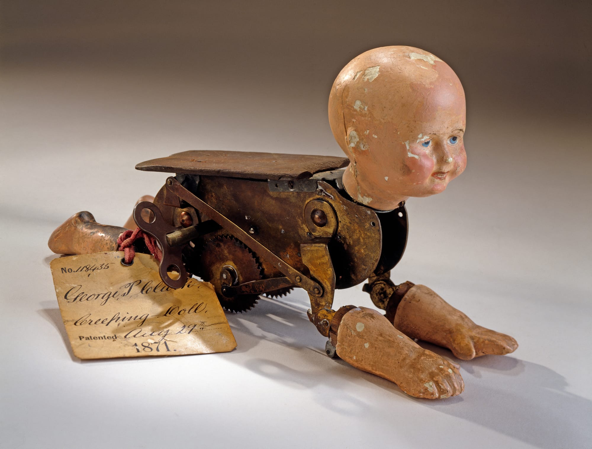 Alt text courtesy Smithsonian Open Access: This model demonstrates the invention of a mechanical crawling doll. It accompanied the patent submission of George Pemberton Clarke, who received U.S. patent No. 118,435 on 29 August 1871 for his “Natural Creeping Baby Doll.” The original patent office tag is still attached with red tape. Clarke’s patent was an improvement on the crawling baby doll patent of his associate Robert J. Clay (No. 112,550 granted 14 March 1871). The doll’s head, two arms and two legs are made of painted plaster. The arms and legs are hinged to a brass clockwork body that actuates the arms and legs in imitation of crawling, but the doll moves forward by rolling along on two toothed wheels. A flat piece of wood is attached to top of the movement.