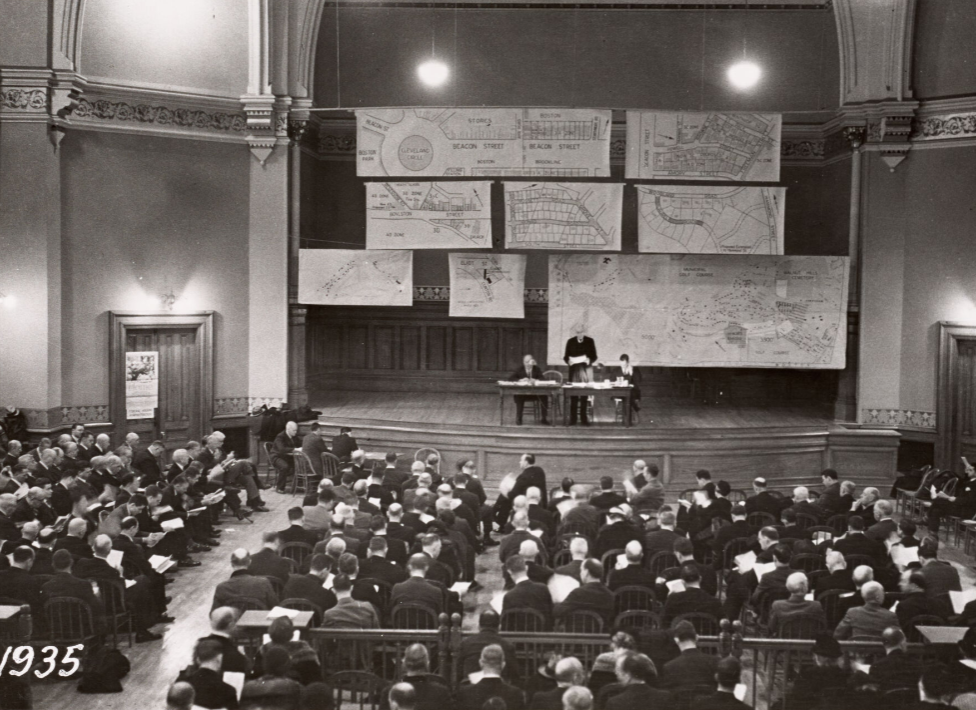 A photograph from a town meeting in 1935. Hundreds of people in an auditorium, three on stage, with a number of maps hung from the rafters.