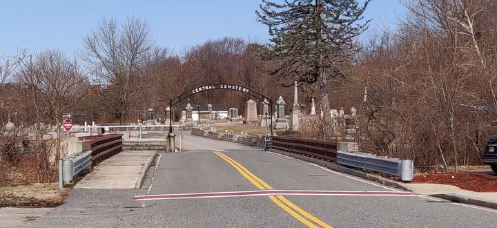 A photo of the road leading to Central Cemetery in Millbury. The road crosses a bridge and under a black metal arch.